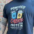 Person wearing a black t-shirt with 'Powered by Caffeine and Protein' design against a brick wall.