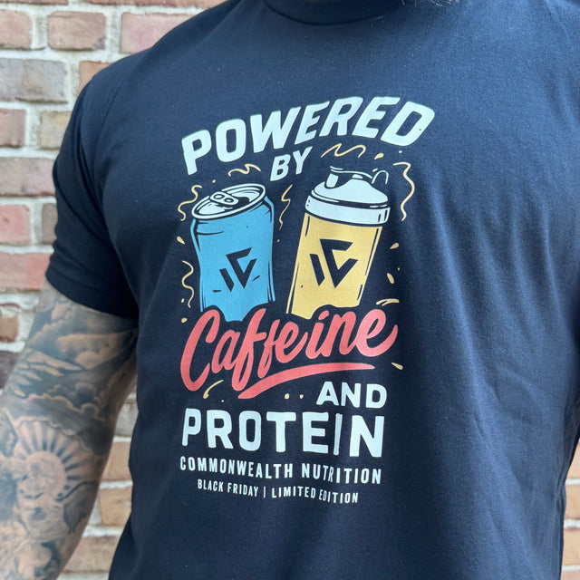 Person wearing a black t-shirt with 'Powered by Caffeine and Protein' design against a brick wall.